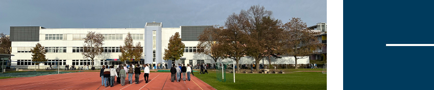 Schulgebäude mit rotem Sportplatz im Vordergrund, davor eine Gruppe von Menschen